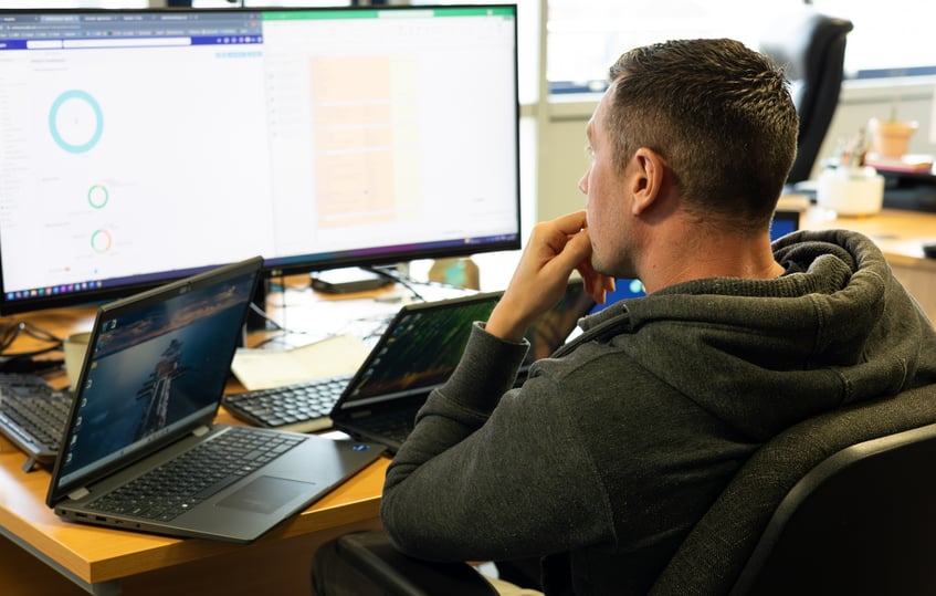 IT technician sitting at a desk looking at a screen showing analytics, as well as two laptops open in front of him